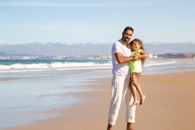 Joyful handsome dad holding happy little daughter in arms, standing wet sand, enjoying leisure time with girl on beach at sea