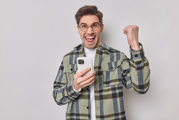 Joyful handsome European man feels glad celebrates winning online game clenches fist with triumph holds mobile phone wears round spectacles and checkered shirt isolated over white background.