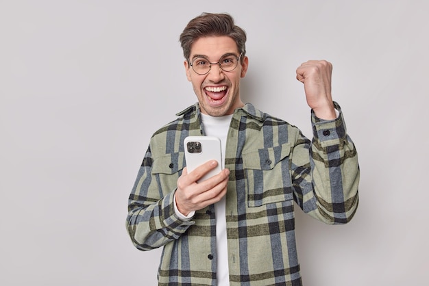 Радостный красивый европейский мужчина чувствует себя счастливым, празднует победу в онлайн-игре, триумфально сжимает кулак, держит мобильный телефон, носит круглые очки и клетчатую рубашку, изолированную на белом фоне.