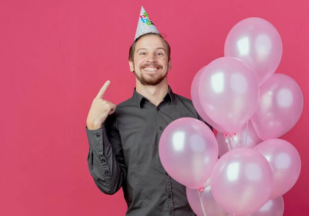 Радостный красавец в кепке дня рождения стоит с гелиевыми шарами, указывающими на кепку, изолированную на розовой стене