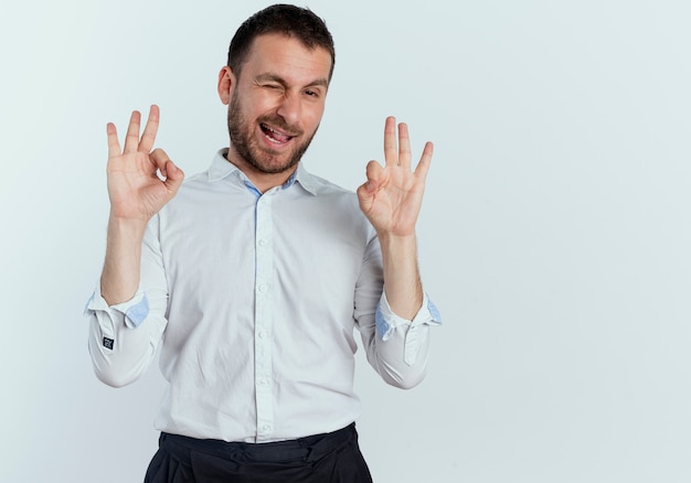 Joyful handsome man blinks eye and gestures ok hand sign with two hands isolated on white wall