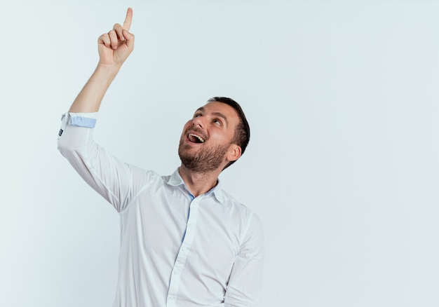 Joyful handsome man points and looks up isolated on white wall