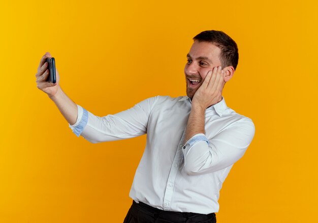 Joyful handsome man puts hand on face looking at phone taking selfie isolated on orange wall