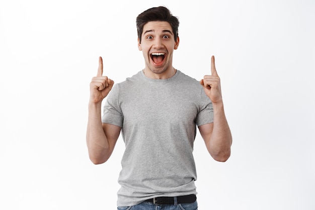 うれしそうなハンサムな男は喜びから叫び、指を上に向け、上部のコピースペースに広告を表示し、白い背景にTシャツで立っています