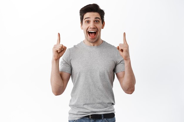즐거운 잘 생긴 남자는 기쁨에서 비명을 지르고 손가락을 위로 가리키며 위쪽 복사 공간에 광고를 표시하고 흰색 배경에 대해 티셔츠를 입고 서 있습니다.