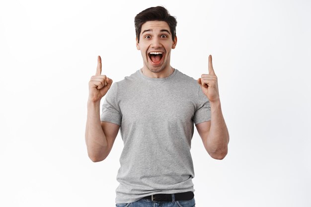 Joyful handsome man scream from joy and pointing fingers up, showing advertisement on top copy space, standing in t-shirt against white background