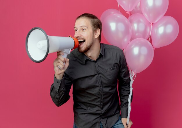 Joyful handsome man stands with helium balloons holding loud speaker looking at side isolated on pink wall