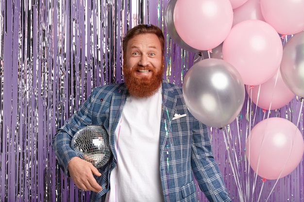 Joyful handsome man throws party after promotion at work, holds disco ball and bunch of air balloons, smiles happily, wears elegant jacket, stands over purple tinsel curtain, enjoys loud music