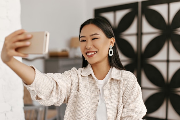 베이지색 재킷을 입은 즐거운 행복한 브루네트 여성은 아늑한 방에서 진심으로 미소 짓는 아시아 여성이 전화를 들고 셀카를 찍는다