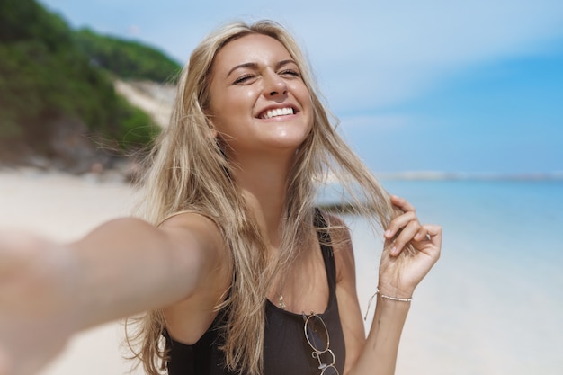 Joyful happy carefree blond tanned woman enjoyings sun, closes her eyes raise and takes selfie in sandy beach