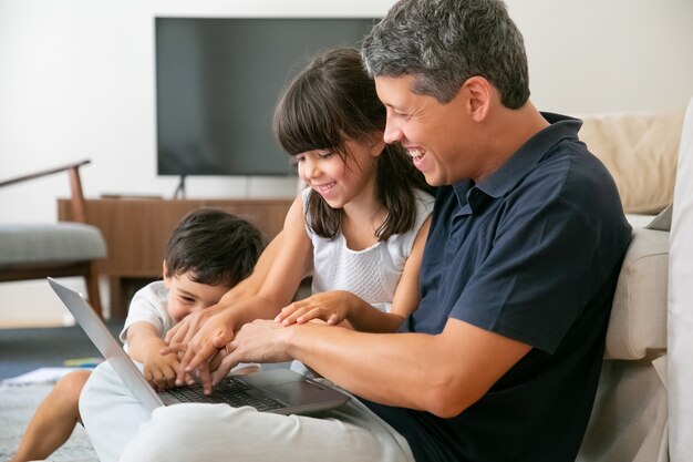 Joyful happy dad and two kids using laptop together, sitting on floor in apartment, pressing buttons in keyboards.