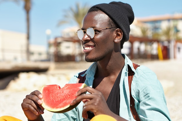 Joyful happy fashionable young Afro American man having picnic on beach with friends, holding slice of ripe watermelon, looking towards sea with broad smile, watching dolphins playing in water