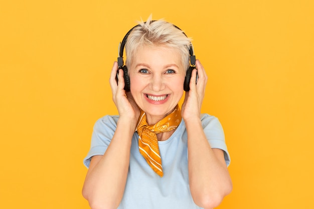 Joyful happy mature short haired woman smiling broadly posing on yellow in wireless headphones
