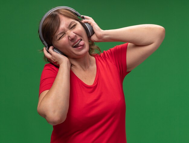 Joyful happy middle aged woman in red t-shirt with headphones enjoying her favorite music sticking out tongue standing over green wall