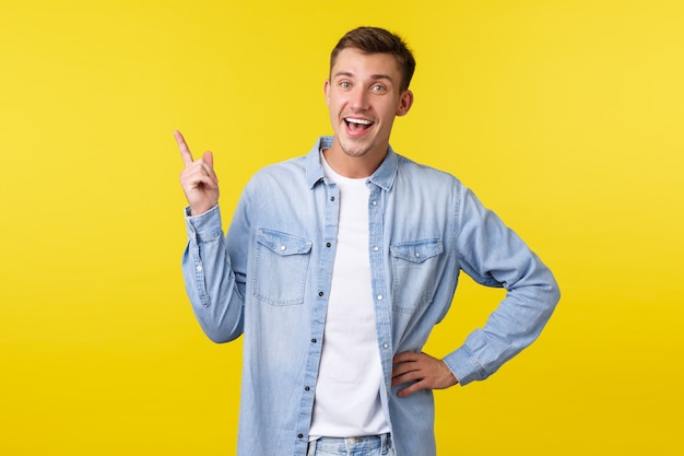 Joyful happy smiling caucasian man pointing finger upper left corner and looking camera delighted. Guy recommend advertisement of product, talking about great new offer, yellow background.