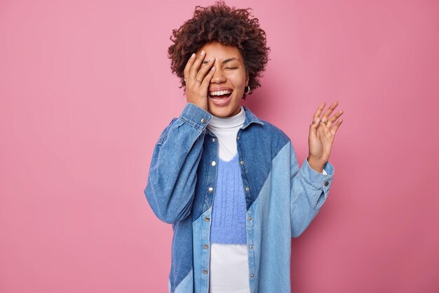Joyful happy woman makes face palm laughs gladfully feels optimistic wears casual jumper and denim shirt smiles broadly isolated over pink background. People authentic emotions feelings concept