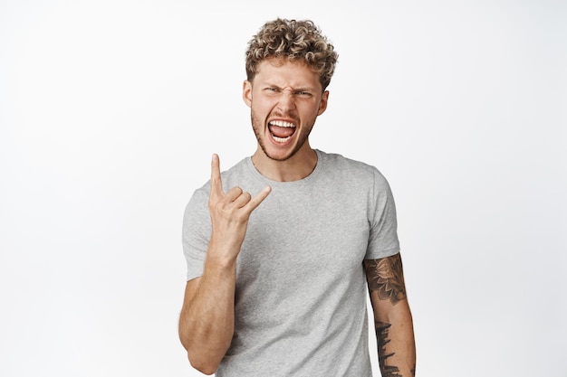 Joyful hipster guy showing rock n roll heavy metal horns gesture shouting excited and enjoying party or event standing over white background