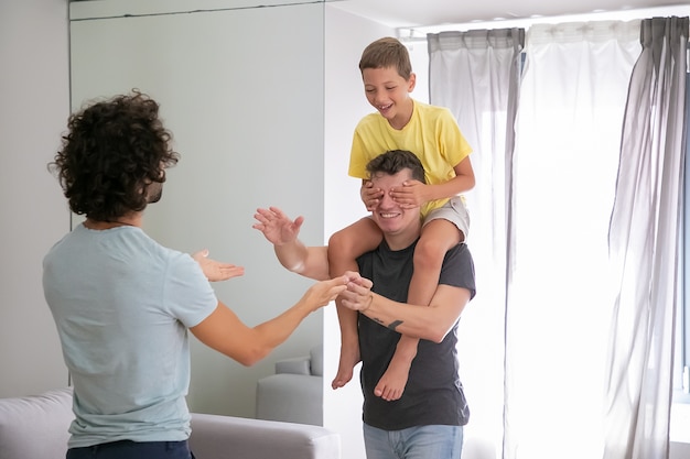 Free photo joyful homosexual dads and his son playing active games at home, having fun. boy riding on mans neck and shutting dads eyes with hands. family and parenthood concept