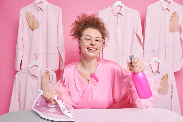 Joyful housewife spends all day on cleaning and ironing uses water spray bottle electric iron wears transparent glasses and dressing gown being at home poses near ironed clothes. Housekeeping