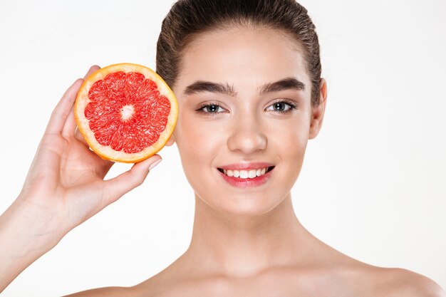 彼女の顔の近くのオレンジ色の柑橘類を押しながら見ている自然なメイクと半分裸の女性を笑顔のうれしそうな画像