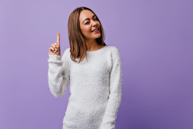 Joyful initiative girl winks, showing that she has new idea. Pretty student happy to arrival of muse and posing on purple wall with space for text