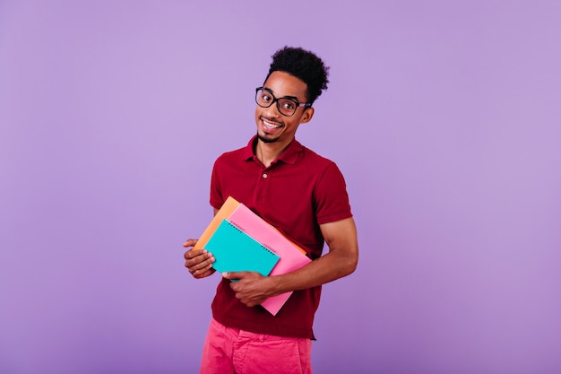 Радостный международный студент в больших очках смотрит. Крытый портрет умного африканца в красной футболке позирует с учебниками.