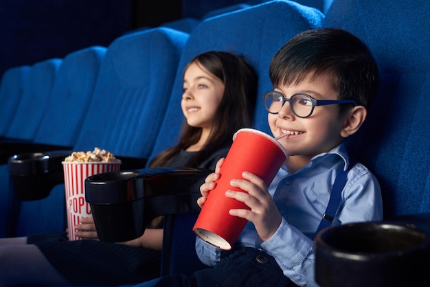 Joyful kids watching movie, drinking fizzy drink in cinema.