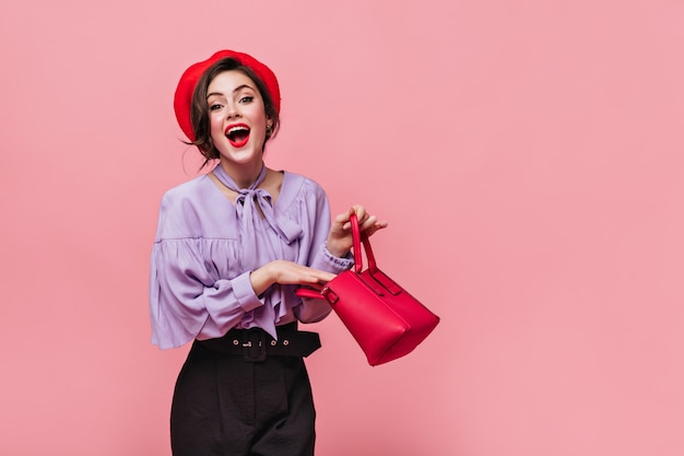 Joyful lady in red hat laughs, holding small bag in her hands on pink background.