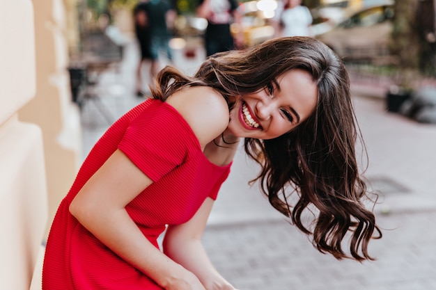 Радостная дама с элегантной прической, выражая счастье по городу. Чувственная брюнетка женская модель в красном наряде смеется на улице.