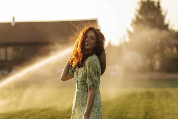 Joyful lady with ginger hairstyle straw hat and black bandage on neck in modern green clothes laughing and looking into camera outdoor