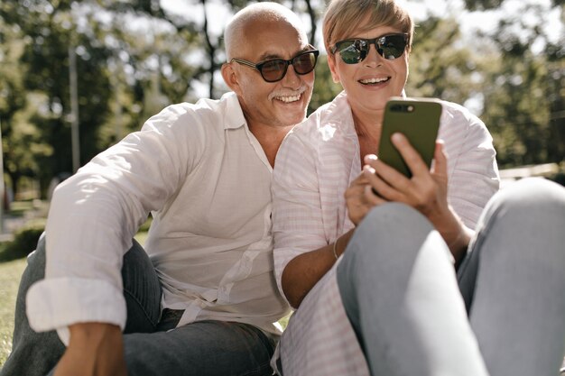 Joyful lady with short hair in sunglasses and striped shirt smiling holding smartphone and opposing with happy old man with mustache in park