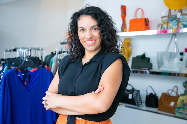 Free photo joyful latin black haired woman standing with arms folded near rack with dresses in clothes shop, looking at camera and smiling. boutique customer or shop assistant concept