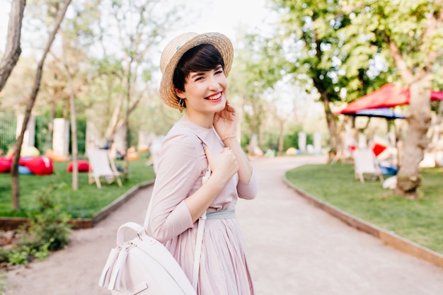 Joyful laughing girl in cute straw hat walking in park in sunny morning, enjoying good weather