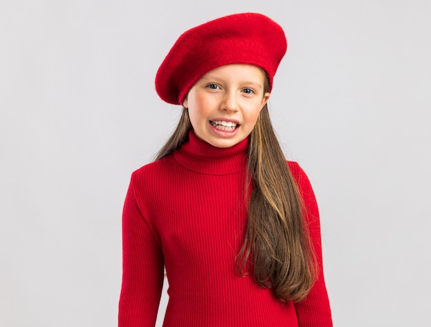 カメラを見て、コピースペースと白い壁に孤立した笑顔の赤いベレー帽を身に着けているうれしそうな小さなブロンドの女の子