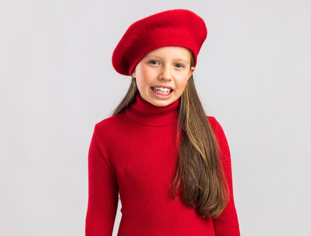 Joyful little blonde girl wearing red beret looking at camera and smiling isolated on white wall with copy space