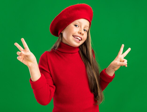 Joyful little blonde girl wearing red beret looking at front showing peace sign isolated on green wall