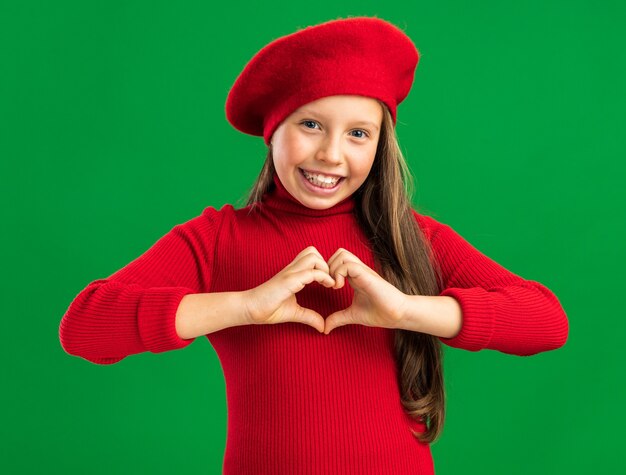 Joyful little blonde girl wearing red beret showing love gesture looking at front isolated on green wall with copy space