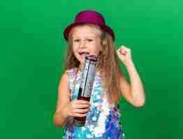 Free photo joyful little blonde girl with purple party hat holding confetti cannon and keeping fist up isolated on green wall with copy space