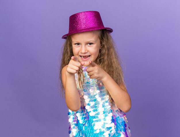 joyful little blonde girl with violet party hat pointing  isolated on purple wall with copy space