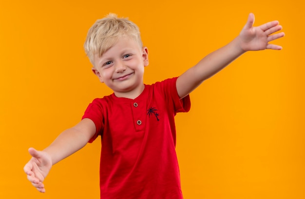 A joyful little boy with blonde hair and blue eyes wearing red t-shirt opening his arms wide for hug while looking on a yellow wall