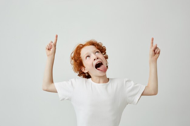 Joyful little boy with curly ginger hair and freckles pointing upside with happy silly face and opened mouth.