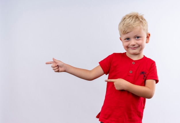 Un gioioso ragazzino biondo carino in maglietta rossa che punta con il dito indice mentre guarda su un muro bianco