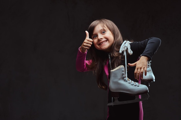 Радостная маленькая девочка, одетая в спортивную одежду, держит коньки и показывает большой палец вверх. Изолированные на темном текстурированном фоне.