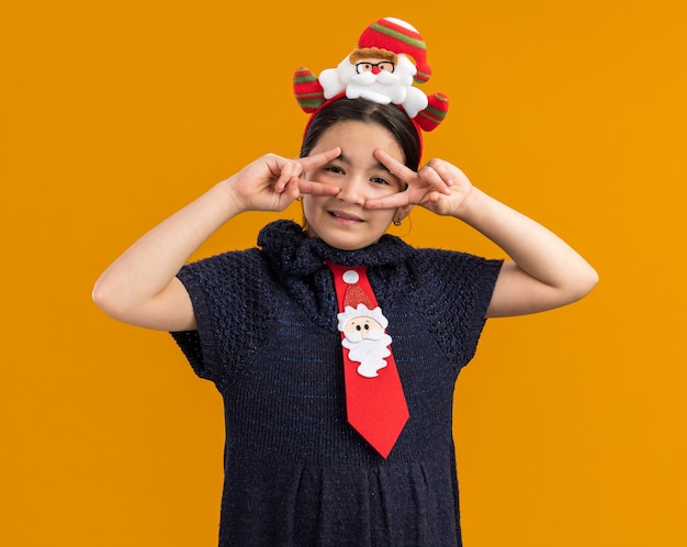 радостная маленькая девочка в вязаном платье в красном галстуке с забавным рождественским ободком на голове смотрит в камеру, показывая знак v рядом с глазами, стоящими на оранжевом фоне