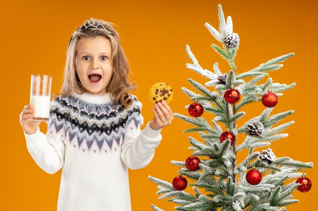 Joyful little girl standing nearby christmas tree wearing tiara with garland on neck holding glass of milk with cookies isolated on orange background