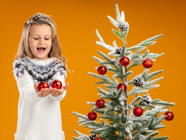 Joyful little girl standing nearby christmas tree wearing tiara with garland on neck holding out christmas balls at camera isolated on orange background