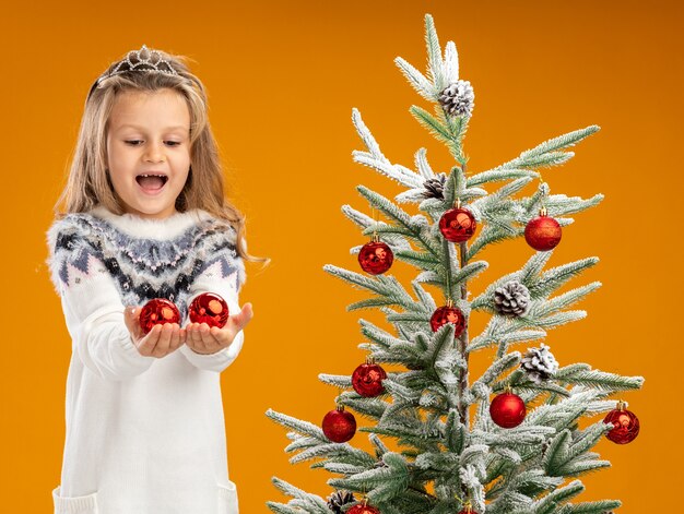 Joyful little girl standing nearby christmas tree wearing tiara with garland on neck holding out christmas balls isolated on orange wall