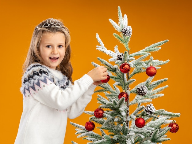 Joyful little girl standing nearby christmas tree wearing tiara with garland on neck holding tree isolated on orange wall