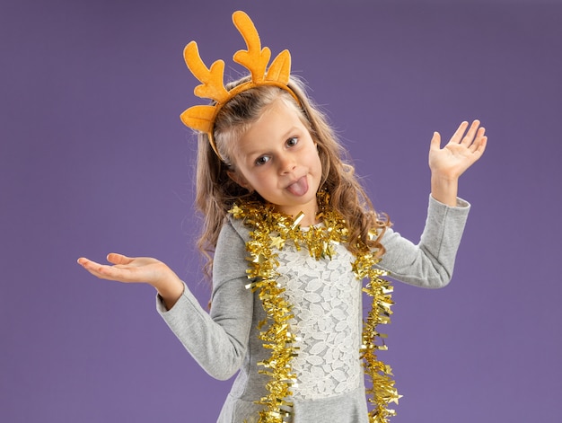 Joyful little girl wearing christmas hair hoop with garland on neck spreading hands and showing tongue isolated on blue background