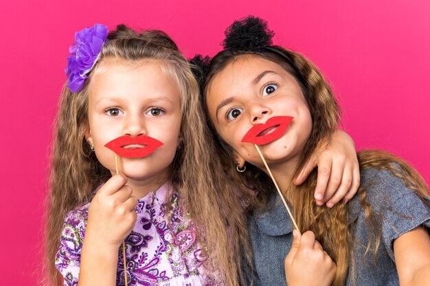 joyful little pretty girls holding fake lips on sticks isolated on pink wall with copy space